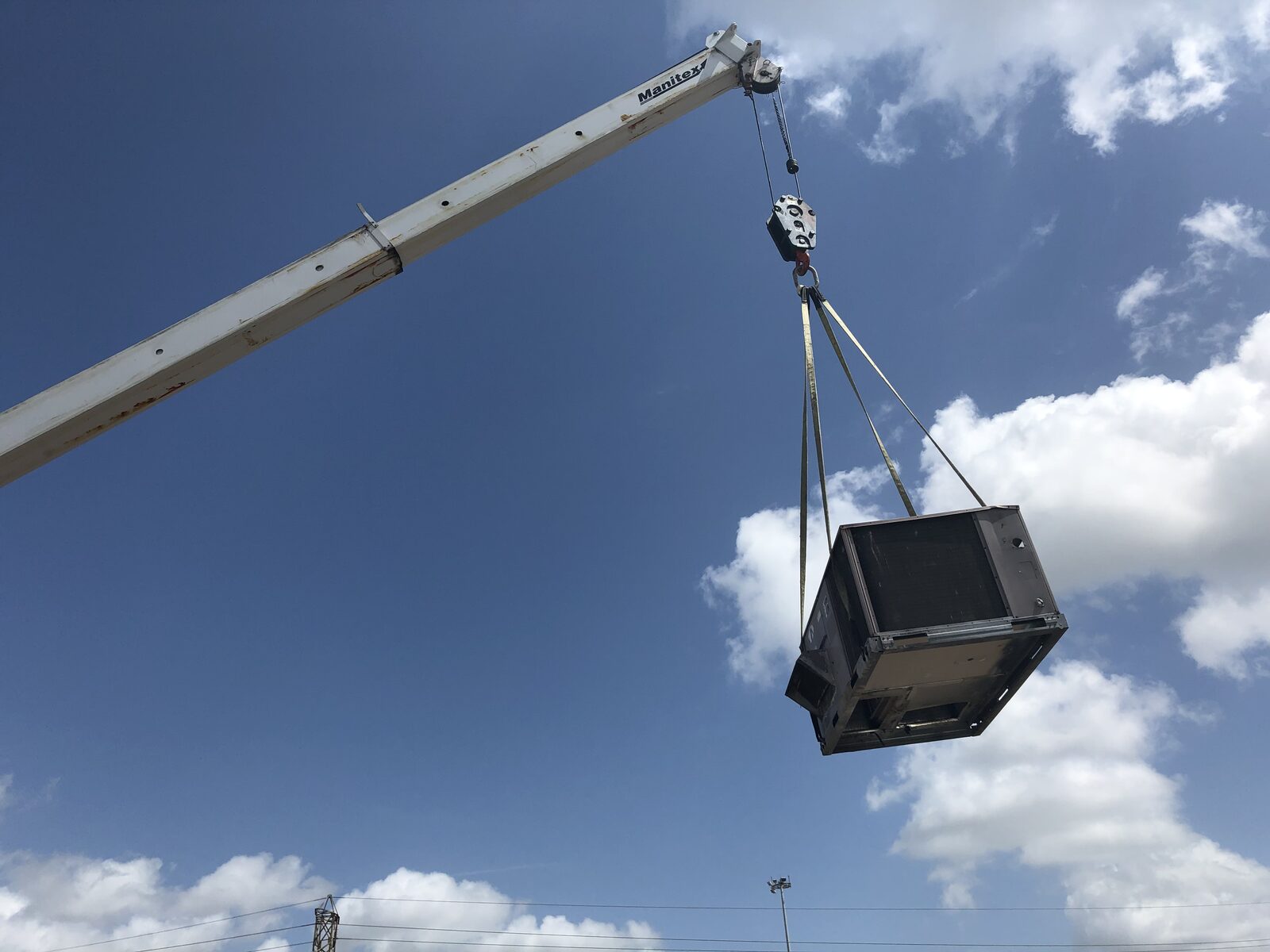 A packaged rooftop air conditioning unit hanging from a crane against a blue sky on a Ferre job